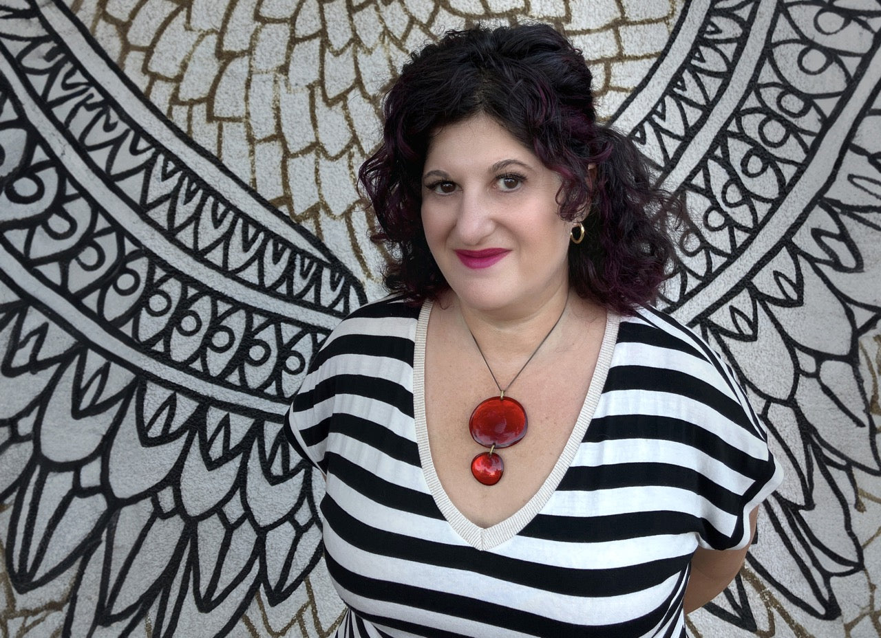 Urban fantasy author Deborah Wilde stands in front of a line drawing mural of wings on the side of a Vancouver building. She is wearing a black and white striped top and a large red pendant necklace.