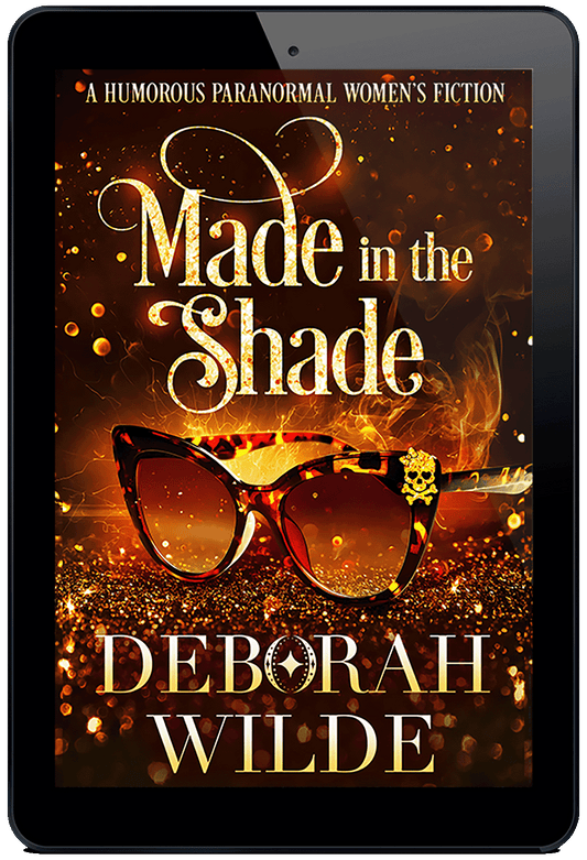 A gold book cover for Made in the Shade by Deborah Wilde. A pair of tortoise shelled sun glasses sits on a field of sparkles with sparks and smoke rising from it. On the top corner of the glasses is a glittery skull with heart shaped eye sockets and a large glittery bow on its head.