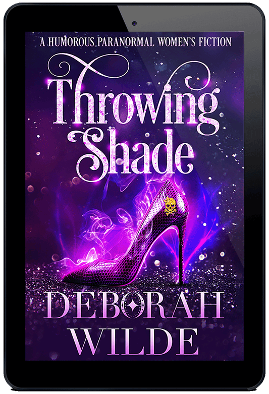 A royal purple book cover for Throwing Shade by Deborah Wilde. A sequinned purple high heeled shoe sits on a field of sparkles with sparks and smoke rising from it. On the shoe is a glittery skull with heart shaped eye sockets and a large glittery bow on its head.